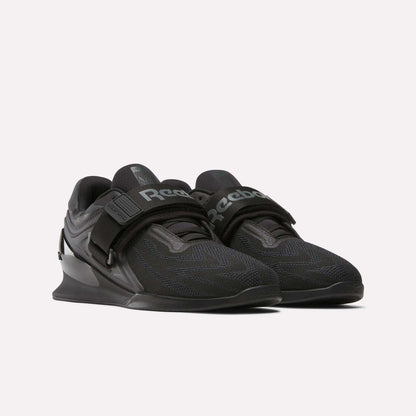 The Reebok Men's Legacy Lifter IV Shoes feature a thick wedge heel, mesh upper, and wide midfoot strap for enhanced stability. Reebok branding is visible on the tongue and strap, shown against a plain white background.
