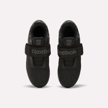 Top view of a pair of Reebok Men's Legacy Lifter IV Shoes in black, with thick textured soles and wide velcro straps for stability. The large Reebok logo is visible. Set against a plain, light-colored background.