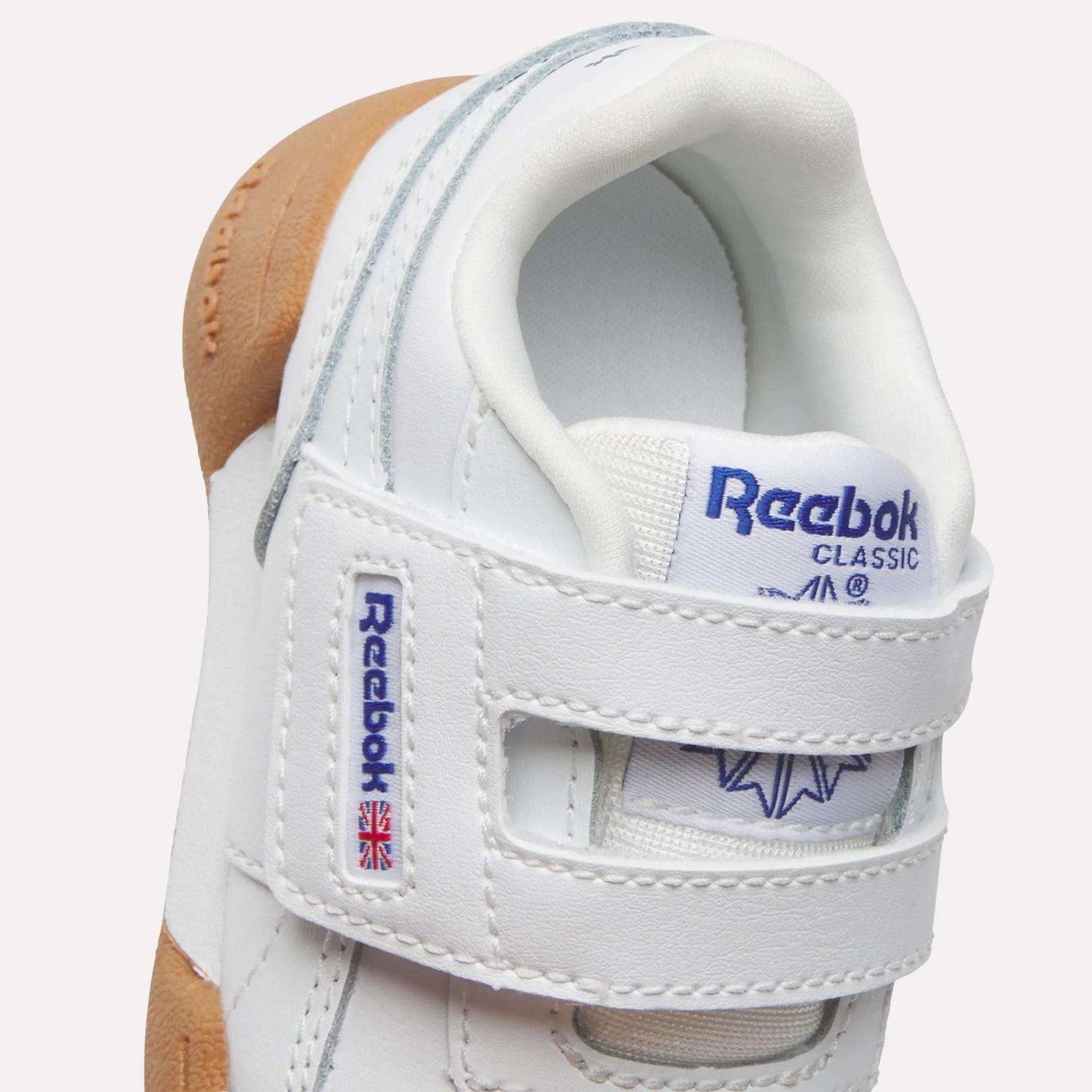 Close-up of Reebok Kids' Workout Plus Shoes for babies and toddlers, featuring white uppers, Velcro straps with brand logo and Union Jack flag, gum-colored sole, and grey accents—ideal for little feet.