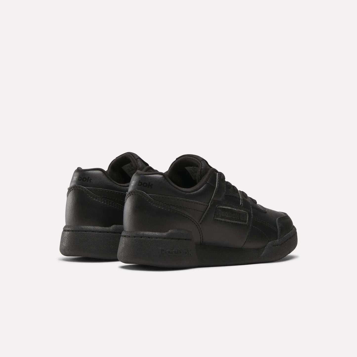 A pair of Reebok Workout Plus Shoes for little kids, shown in black from the back and side. These low-top sneakers feature laces and Reebok branding on the heel and side, displayed against a plain white background.