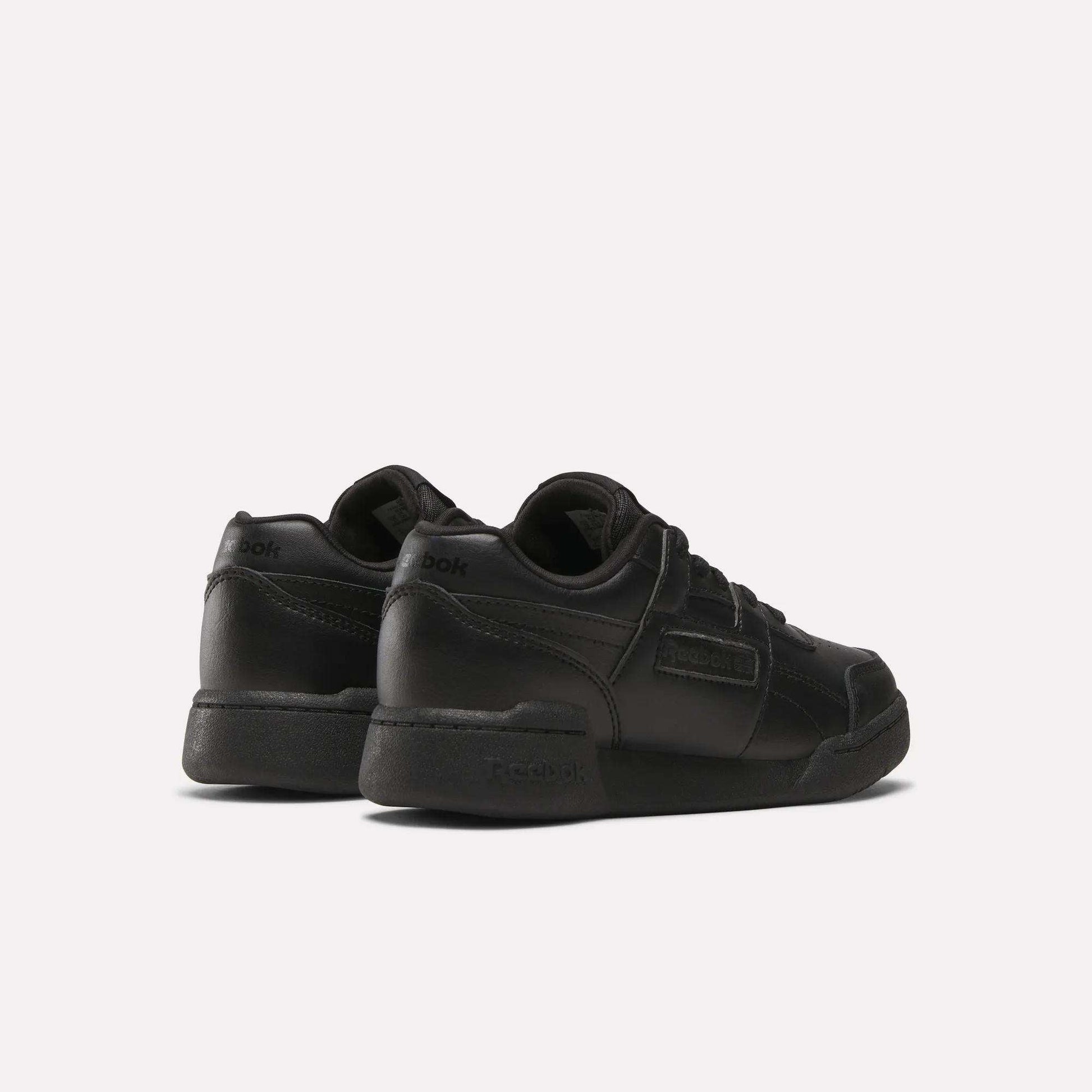 A pair of Reebok Workout Plus Shoes for little kids, shown in black from the back and side. These low-top sneakers feature laces and Reebok branding on the heel and side, displayed against a plain white background.