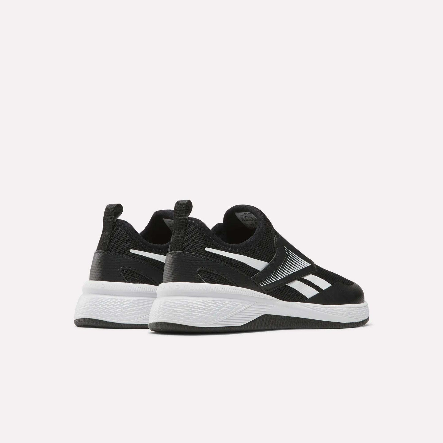 A pair of Reebok Nano Play Slip-On Shoes for little kids in black, with white soles and logo accents, shown facing away and slightly to the side on a plain light background.