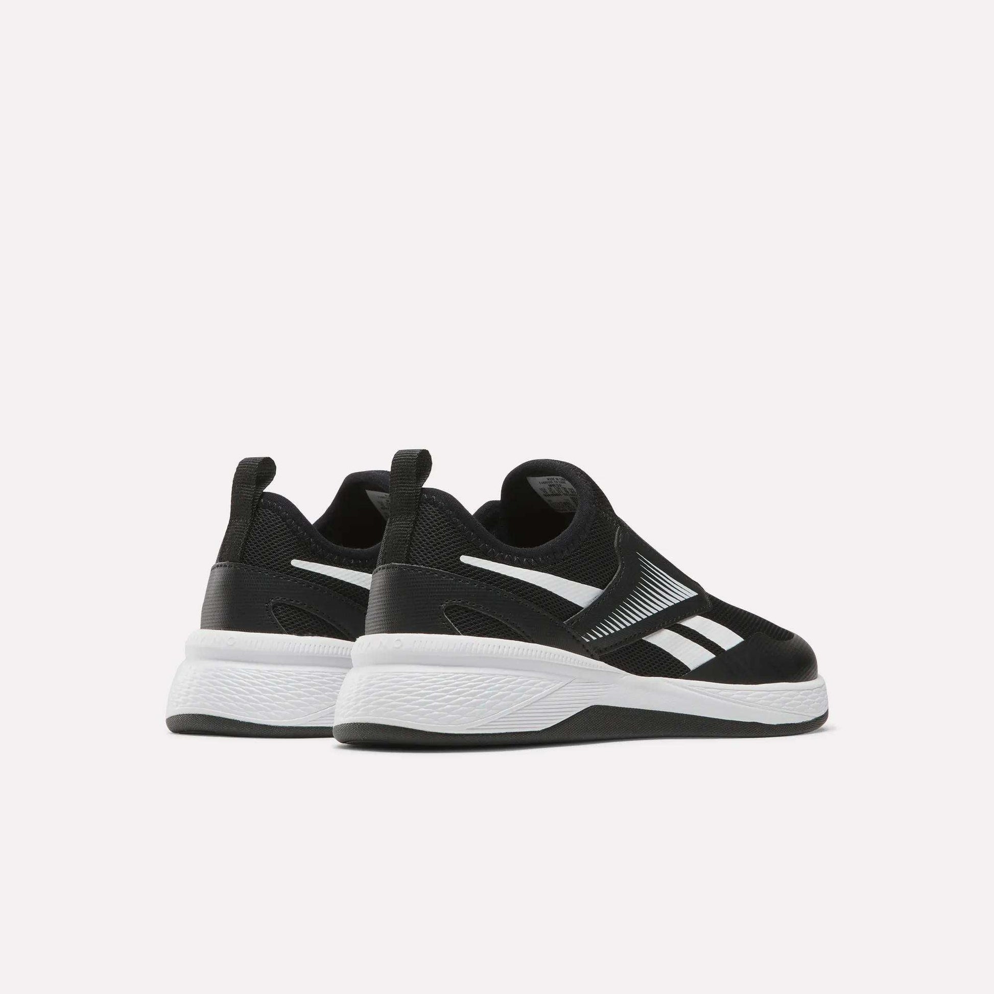 A pair of Reebok Nano Play Slip-On Shoes for little kids in black, with white soles and logo accents, shown facing away and slightly to the side on a plain light background.