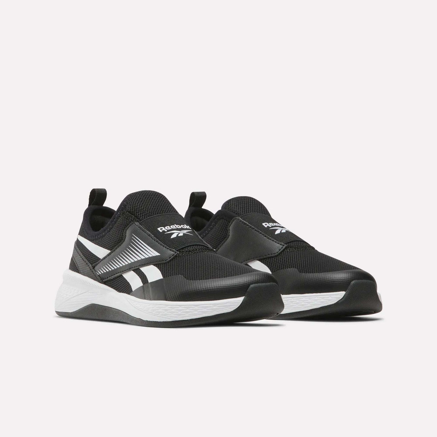 The Reebok Nano Play Slip-On Shoes for little kids, in black with white soles and branding, are low-top athletic shoes featuring heel pull tabs for easy wear, shown on a plain light background.
