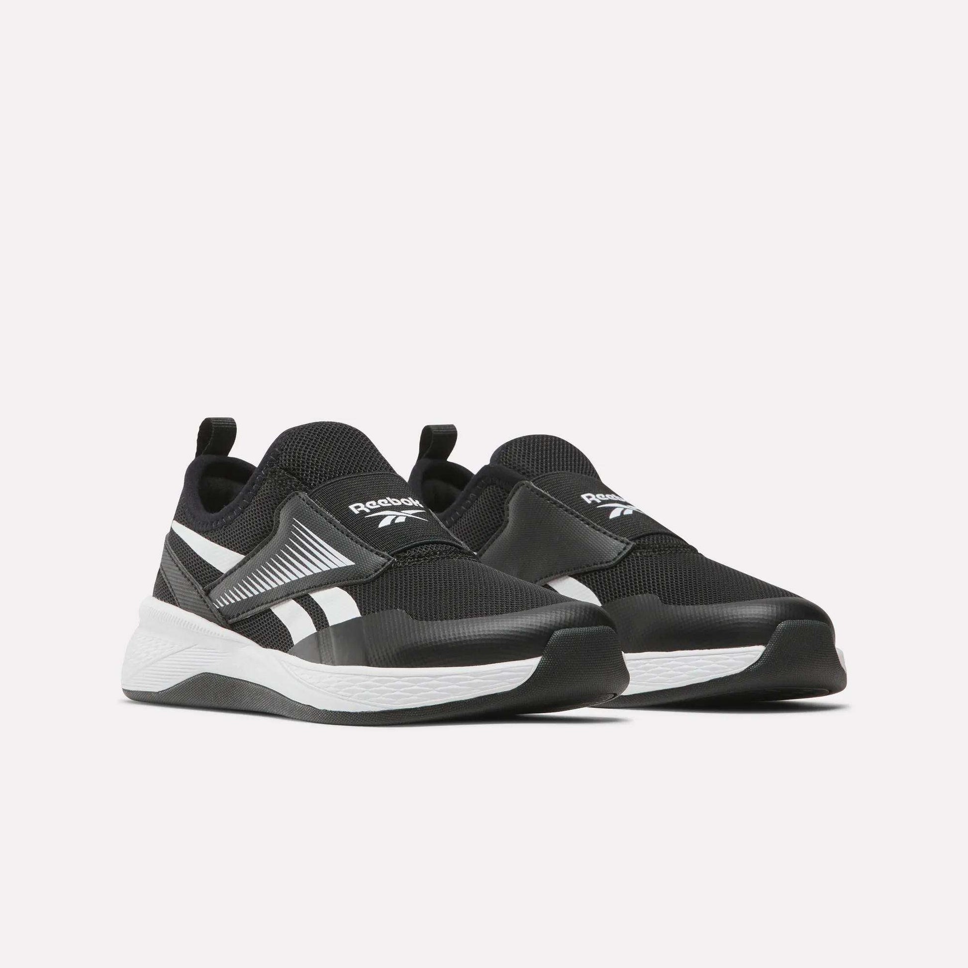The Reebok Nano Play Slip-On Shoes for little kids, in black with white soles and branding, are low-top athletic shoes featuring heel pull tabs for easy wear, shown on a plain light background.