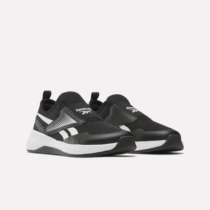The Reebok Nano Play Slip-On Shoes for little kids, in black with white soles and branding, are low-top athletic shoes featuring heel pull tabs for easy wear, shown on a plain light background.