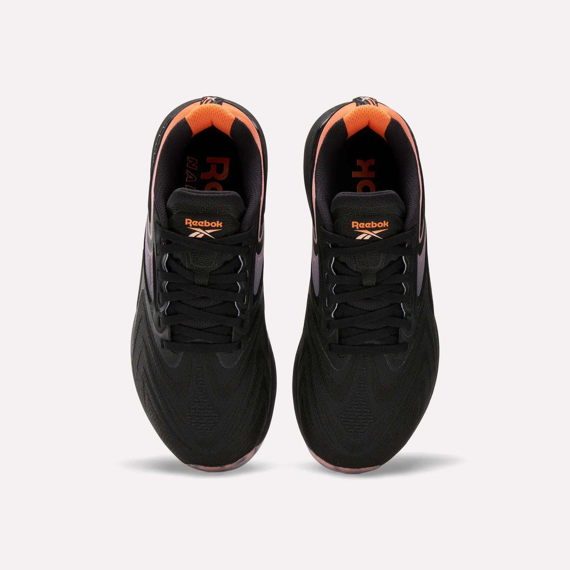 Top view of Women's Nano X5 Edge Training Shoes by Reebok in black with orange heel and inner accents, Reebok logo on tongue and insole. Shoes shown side by side on a white background.