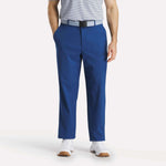 A man stands with his hands in his pockets, wearing a light gray striped polo, white sneakers, and Reebok Men's The Favorite Golf Pants - Relaxed Fit in blue with a black and gray belt against a plain background.