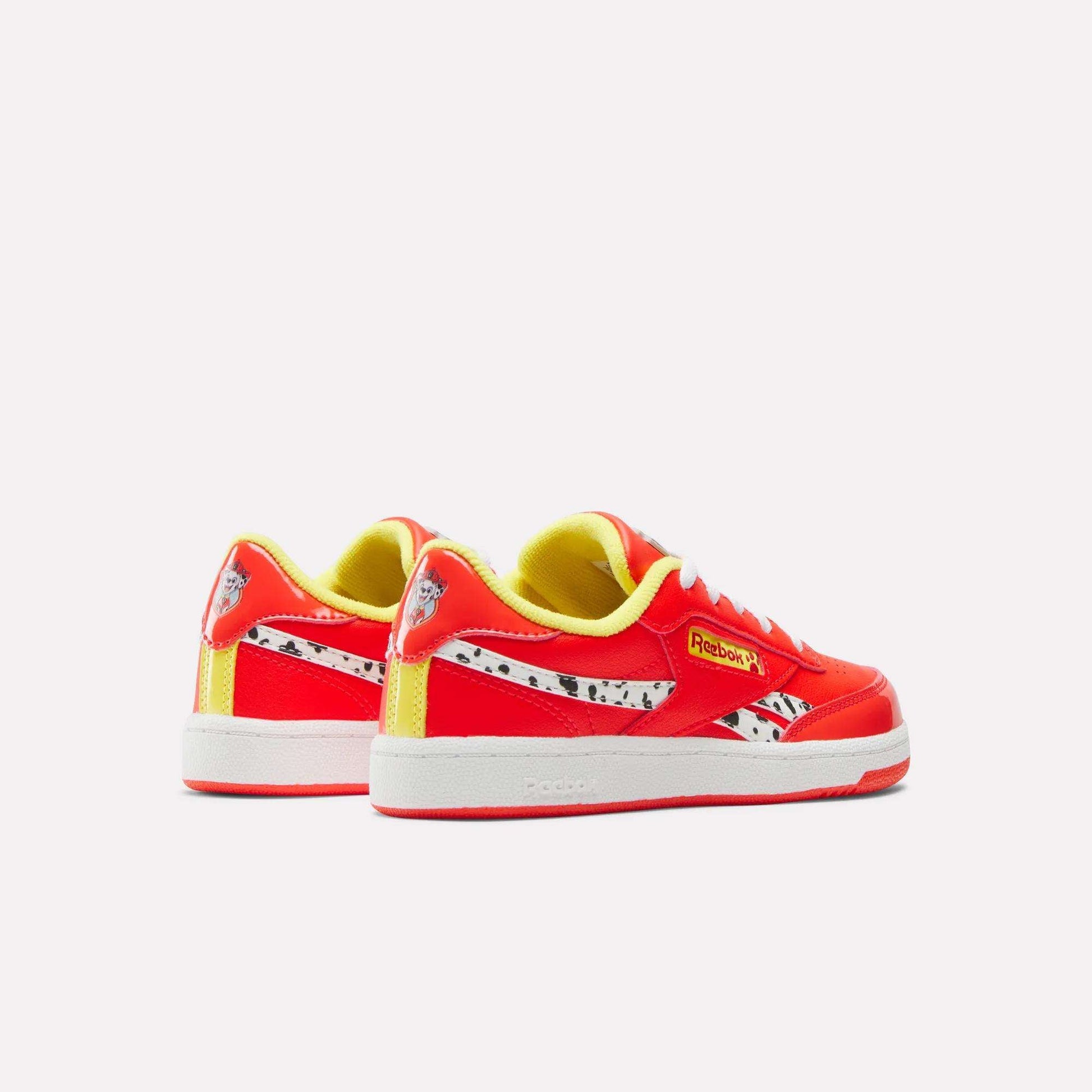 The Reebok x Paw Patrol Club C Revenge Shoes for little kids feature vibrant red and yellow colors, white soles, black spotted accents, and the Reebok logo inspired by retro tennis style, displayed against a plain light background.