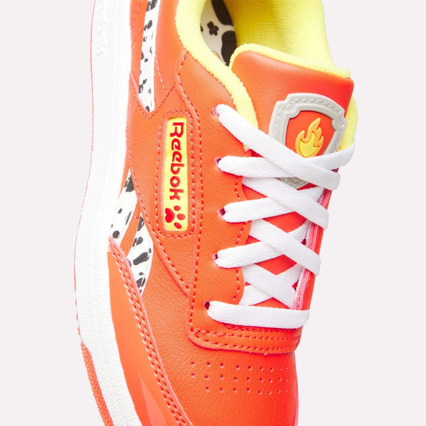 Close-up of the vibrant Reebok x Paw Patrol Club C Revenge Shoes for little kids, featuring orange color, white laces, yellow interior, retro tennis style, black-and-white detail, flame icon on the tongue, and bold red-yellow Reebok logo.