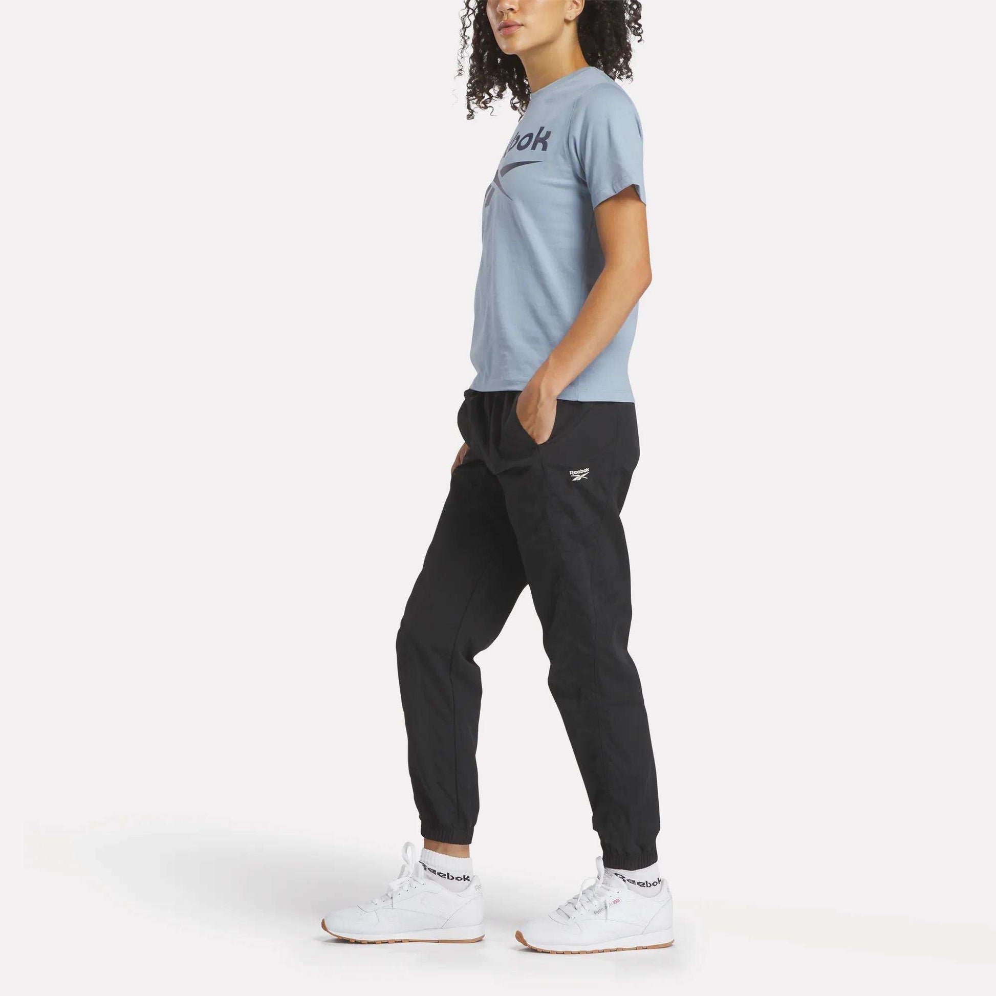 A person with curly hair stands sideways, wearing a light blue Reebok Women's Big Logo Crewneck Short Sleeve Tee, black jogger pants, and white Reebok sneakers against a plain white background.