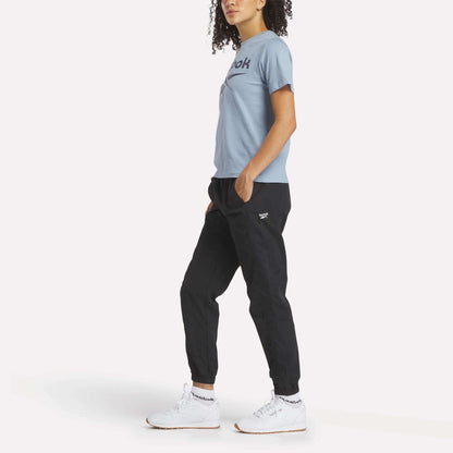 A person with curly hair stands sideways, wearing a light blue Reebok Women's Big Logo Crewneck Short Sleeve Tee, black jogger pants, and white Reebok sneakers against a plain white background.