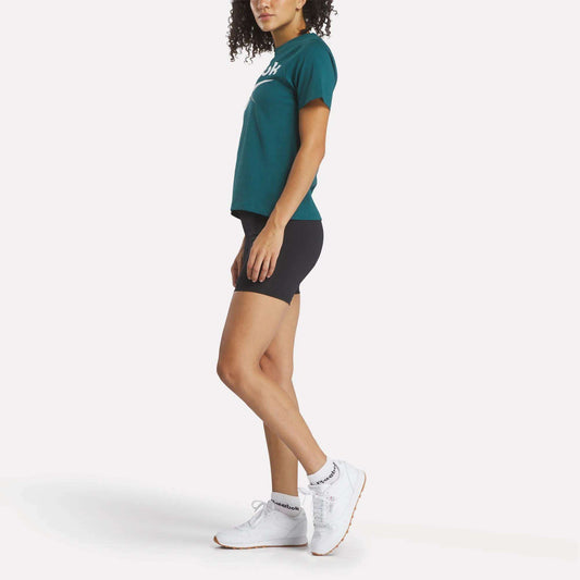 A woman stands in profile view wearing a Reebok Women's Two-Color Logo Crew Short Sleeve Tee with black shorts, white crew socks, and white sneakers against a plain white background.