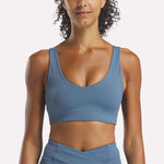 A woman with curly hair models the Reebok Women's Yoga V-Neck Bra and matching high-waisted leggings in blue, shown from shoulders to hips against a plain white background—ideal for studio workouts.