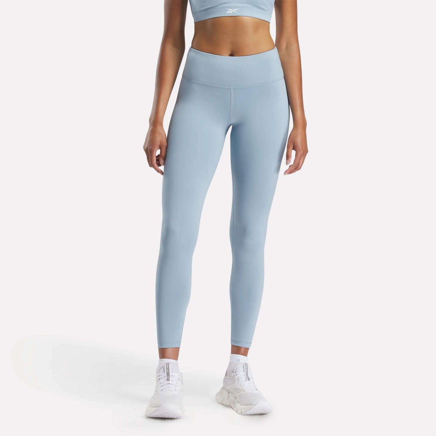 A person wears Reebok Women's Core High-Rise Leggings in light blue, paired with a matching sports bra and white athletic shoes, standing against a plain white background. The cropped photo shows the model from shoulders to feet.