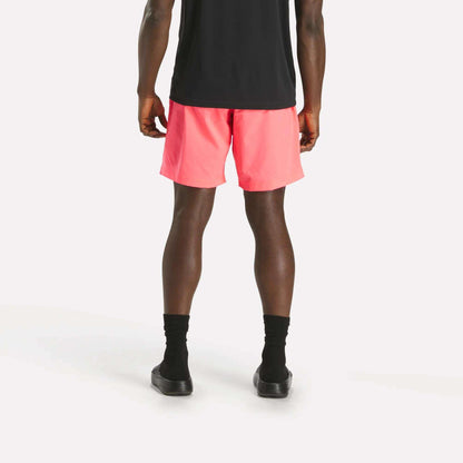 A person faces away from the camera, wearing a black shirt, black socks and shoes, and bright pink Reebok Men's Workout Ready Woven Shorts against a plain white background.