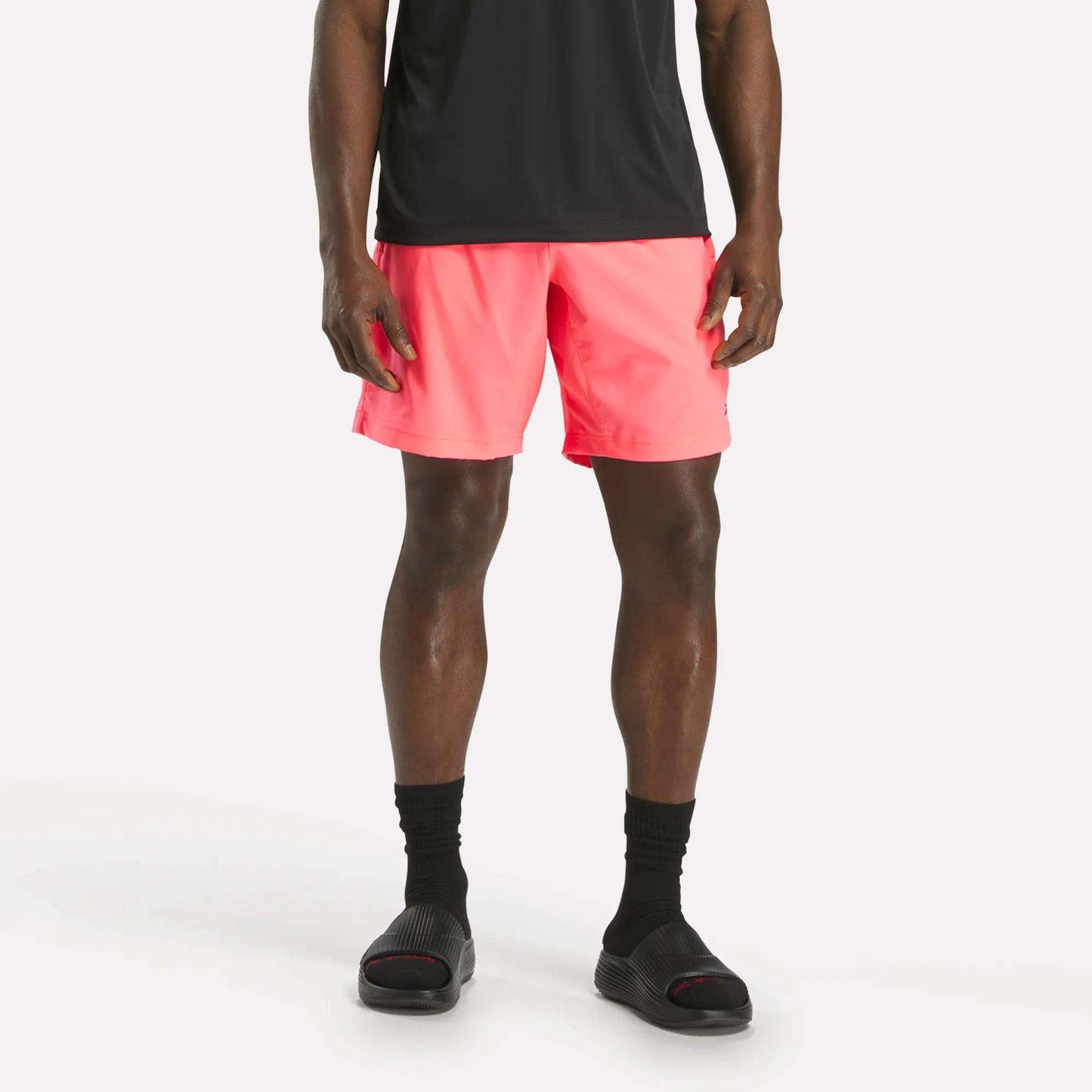A person wearing a black t-shirt, Reebok Men's Workout Ready Woven Shorts in bright pink, black socks, and black slide sandals stands against a plain white background.