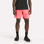 A person wearing a black t-shirt, Reebok Men's Workout Ready Woven Shorts in bright pink, black socks, and black slide sandals stands against a plain white background.