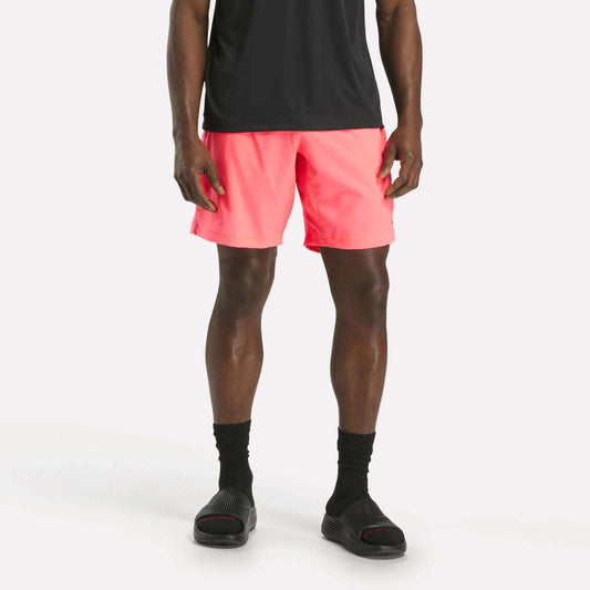 A person wearing a black t-shirt, Reebok Men's Workout Ready Woven Shorts in bright pink, black socks, and black slide sandals stands against a plain white background.