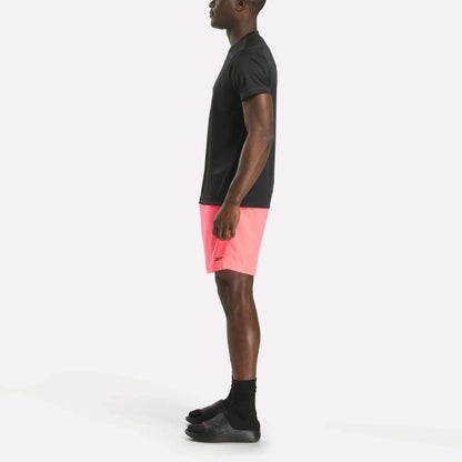 A person stands in profile against a plain white background wearing a black t-shirt, black socks, black slide sandals, and bright pink Reebok Men's Workout Ready Woven Shorts.