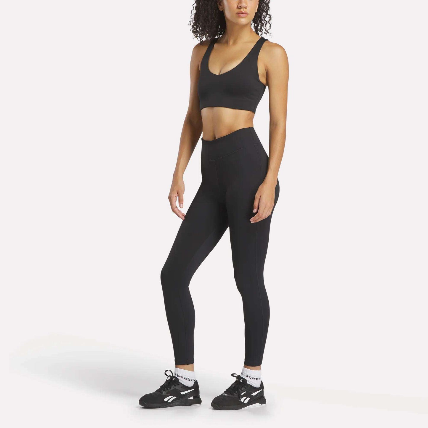 A woman faces forward in Reebok Women's Lux High-Rise Tights, a black sports bra, white socks, and black athletic shoes against a plain white background.