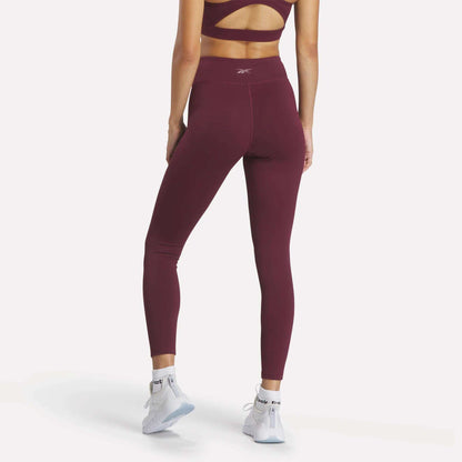 A woman faces away, showcasing the back of her burgundy Reebok Women's Lux High-Rise Tights and matching sports bra, paired with white athletic shoes against a light background.