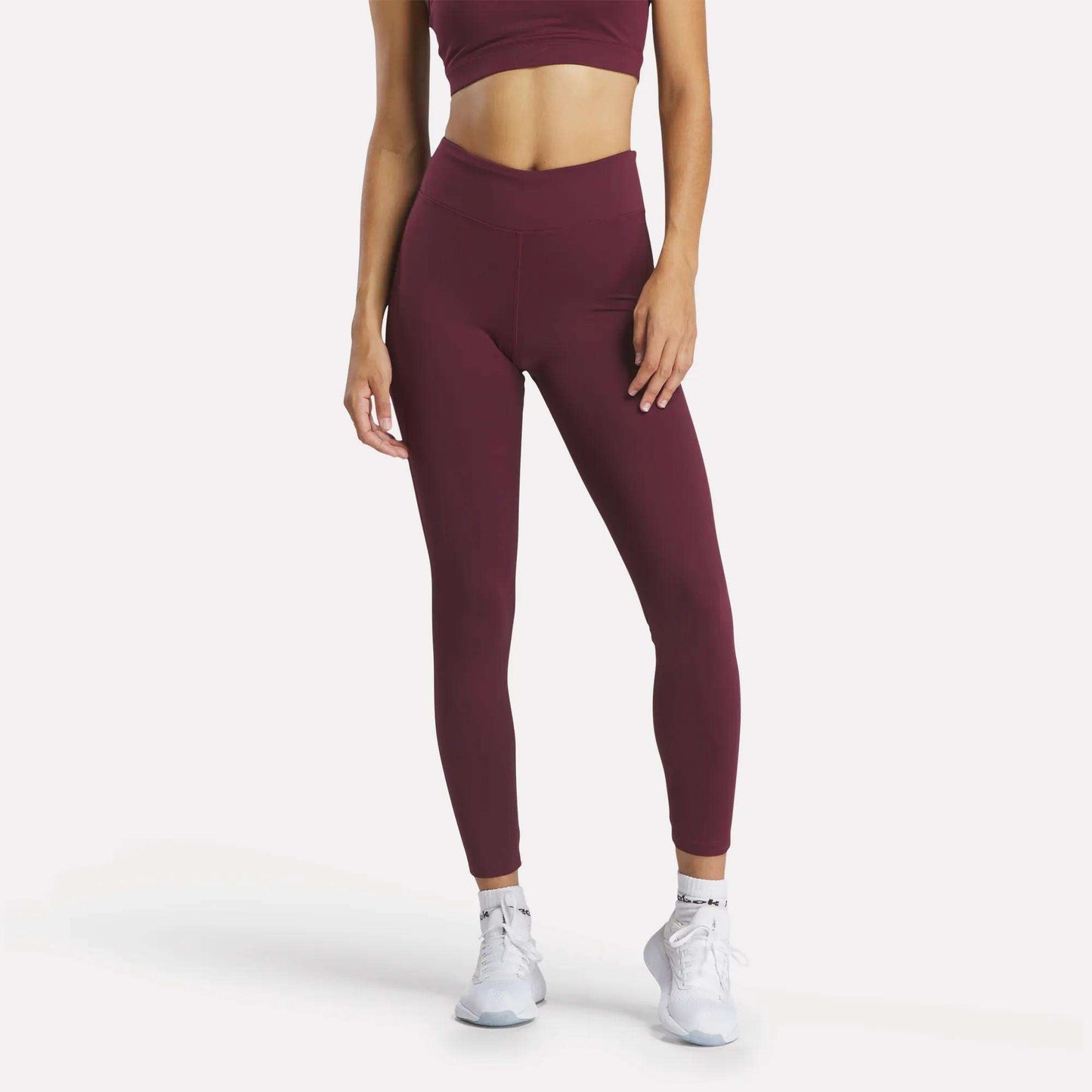A woman models Reebok Women's Lux High-Rise Tights with a burgundy sports bra and white athletic shoes against a white background. Her head is not visible in the image.