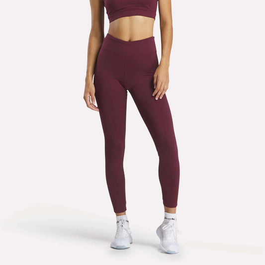 A woman models Reebok Women's Lux High-Rise Tights with a burgundy sports bra and white athletic shoes against a white background. Her head is not visible in the image.