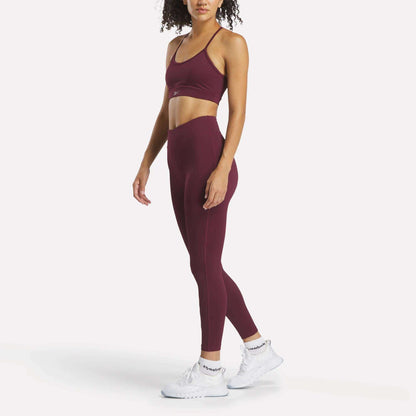 A woman models Reebok Women's Lux High-Rise Tights and a matching burgundy sports bra with RBK-DRY fabric, paired with white athletic shoes. She stands against a white background, slightly turned to the side with relaxed arms.
