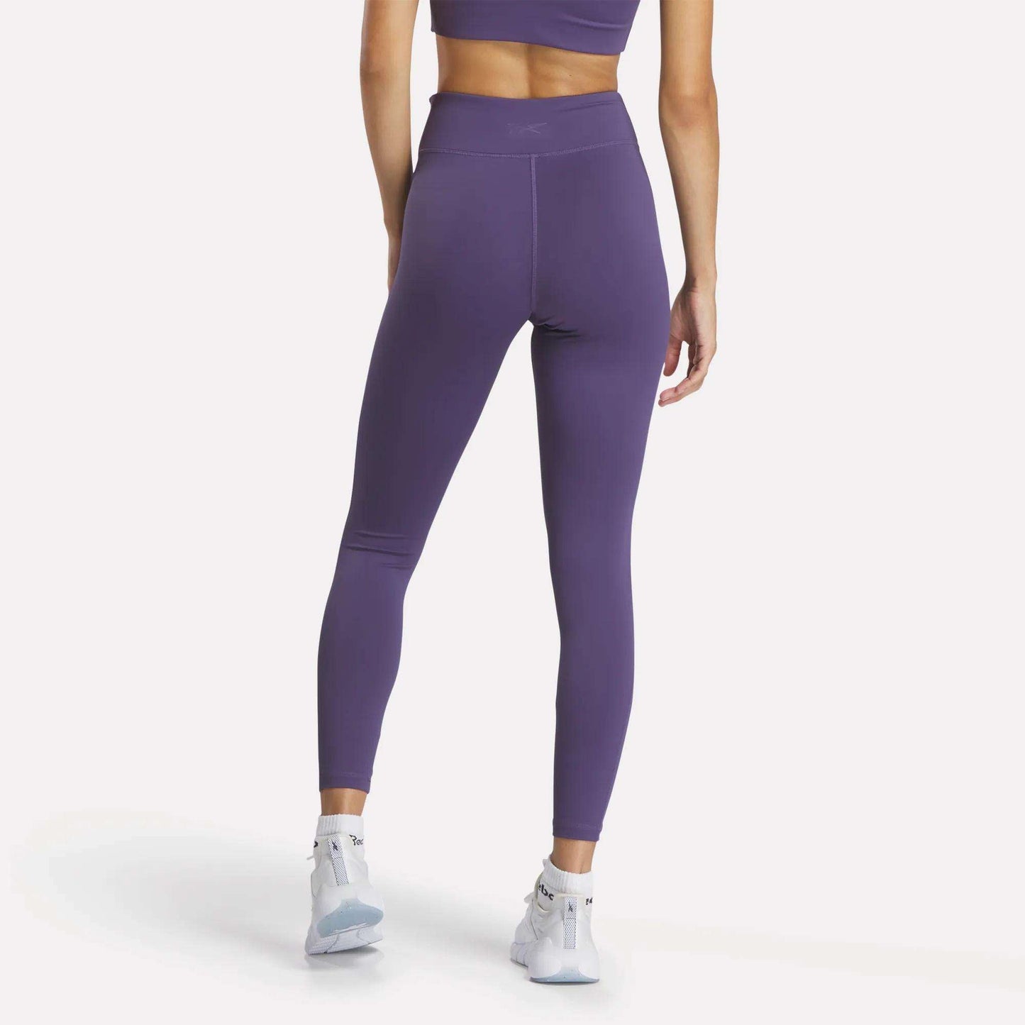 A woman faces away, wearing Reebok Women's Lux High-Rise Tights and a matching purple sports bra with RBK-DRY fabric, plus white athletic shoes, all pictured on a plain white background.