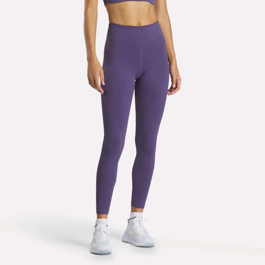 A woman models Reebok Women's Lux High-Rise Tights and a coordinating purple sports bra with white athletic shoes, standing against a plain white background. Her face is not visible.