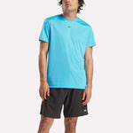 A person wearing a bright blue Reebok Men's ID Train Short Sleeve Tech Tee and black shorts with a small Reebok logo stands against a plain light background.