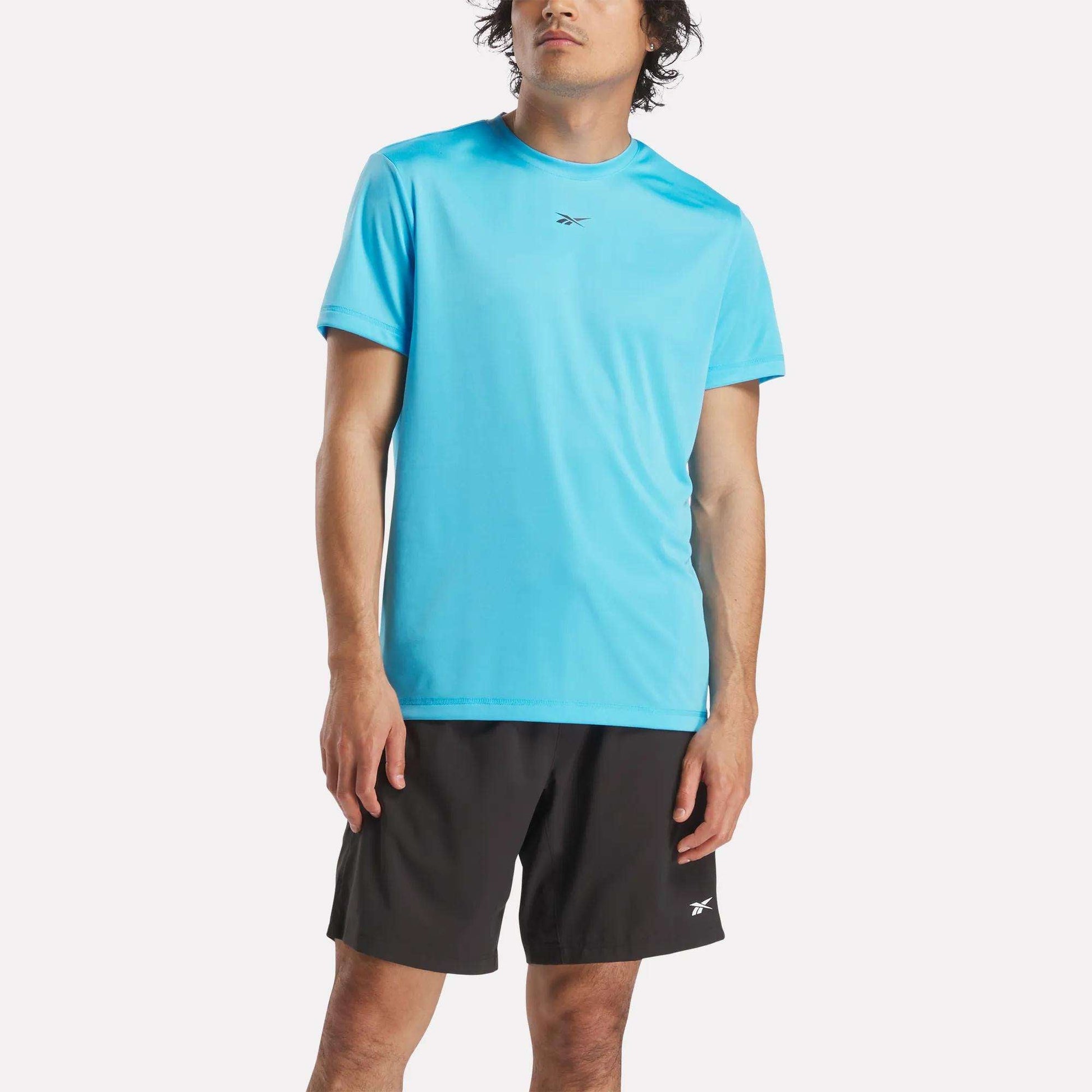 A person wearing a bright blue Reebok Men's ID Train Short Sleeve Tech Tee and black shorts with a small Reebok logo stands against a plain light background.
