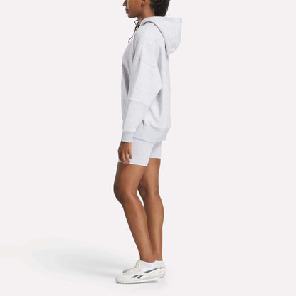 A person stands in profile against a plain white background, wearing a cozy Reebok Women's Lux Hoodie, matching gray shorts, and white sneakers.