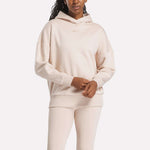 A woman in a light beige Reebok Women's Lux Hoodie and matching leggings stands with her hands clasped. The hoodie features a subtle chest logo, set against a plain white background.