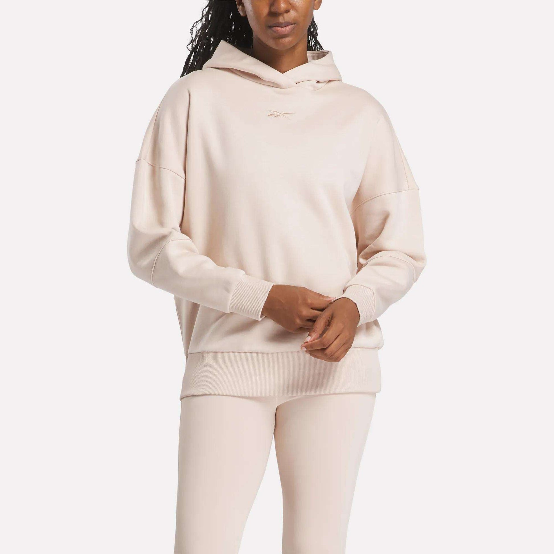 A woman in a light beige Reebok Women's Lux Hoodie and matching leggings stands with her hands clasped. The hoodie features a subtle chest logo, set against a plain white background.