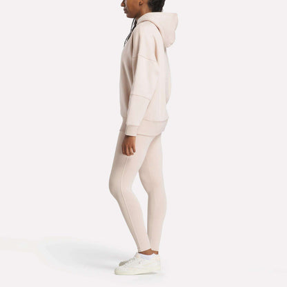 A person stands sideways against a white background, wearing a cozy Reebok Women's Lux Hoodie in beige, matching leggings, and white sneakers.