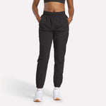 A person wearing a black sports bra, Reebok Women's Vector Track Pants with hands in pockets, and white sneakers featuring orange soles stands against a plain white background.