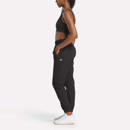 A woman wears Reebok Women's Vector Track Pants in black, paired with a sports bra and white sneakers, standing in profile against a plain white background with her hands in her pockets.