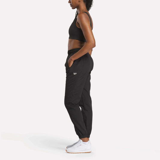 A woman wears Reebok Women's Vector Track Pants in black, paired with a sports bra and white sneakers, standing in profile against a plain white background with her hands in her pockets.