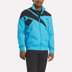 A man models the Reebok Men's Classics Track Top in bright blue with black geometric chest and shoulder patterns, paired with black pants, standing against a plain light background to showcase casual comfort.