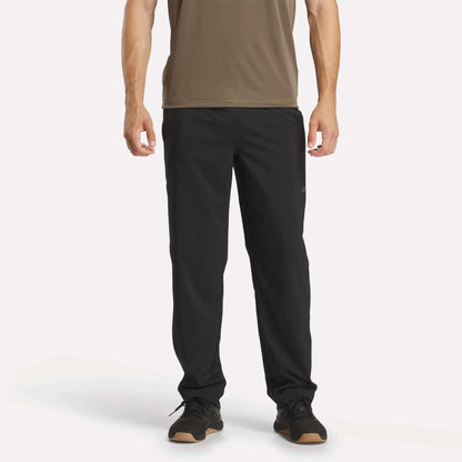 A person wearing a brown short-sleeve shirt, Reebok Men's Train Woven Unlined Pants, and black sneakers stands facing forward against a plain white background.