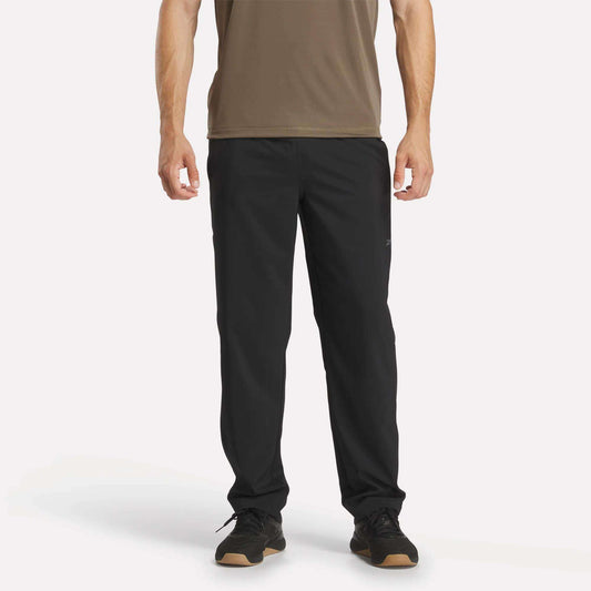 A person wearing a brown short-sleeve shirt, Reebok Men's Train Woven Unlined Pants, and black sneakers stands facing forward against a plain white background.
