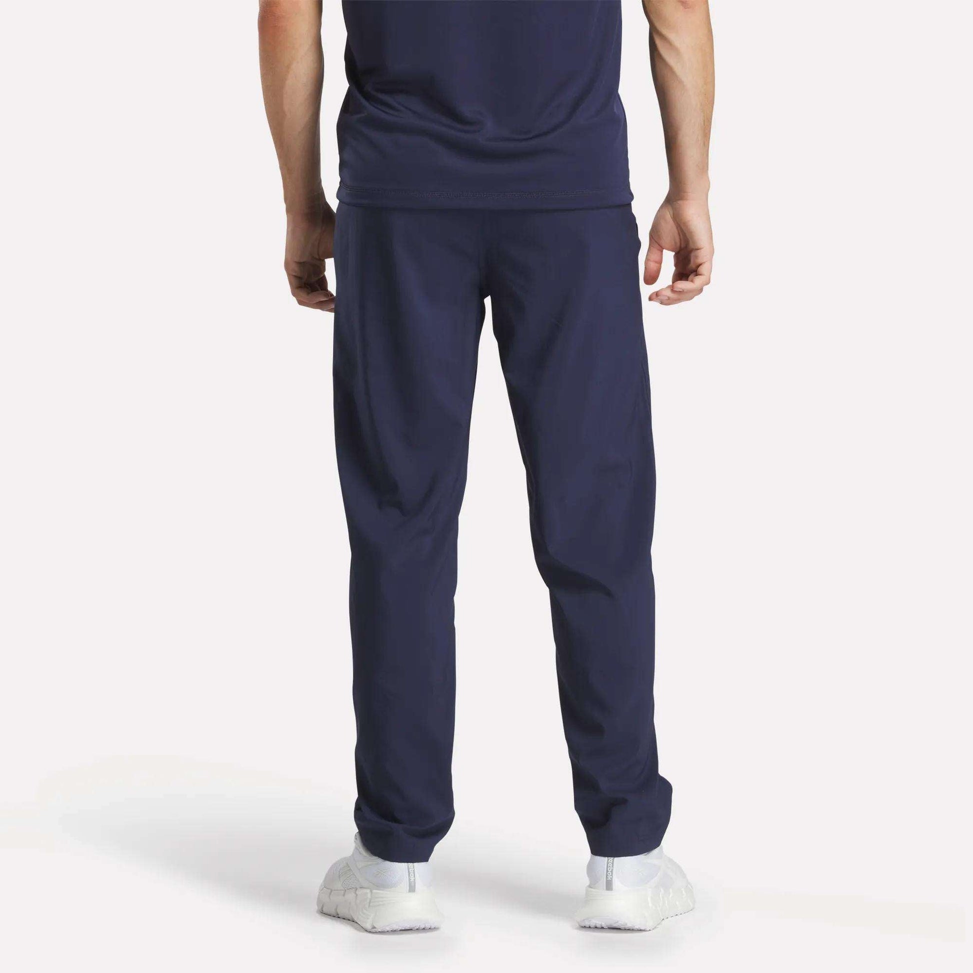 A person faces away, wearing Reebok Men's Train Woven Unlined Pants, a navy blue T-shirt, and white sneakers against a plain light background.