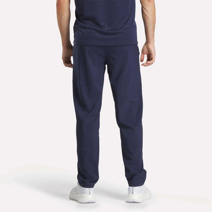 A person faces away, wearing Reebok Men's Train Woven Unlined Pants, a navy blue T-shirt, and white sneakers against a plain light background.