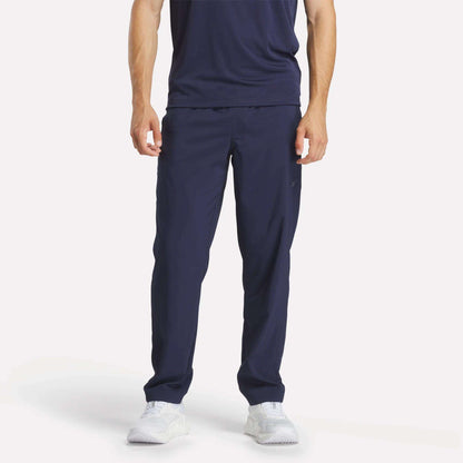 A person stands facing forward against a plain light background, wearing Reebok Men's Train Woven Unlined Pants in navy blue with a matching t-shirt and white athletic shoes. The person's head is not visible in the image.