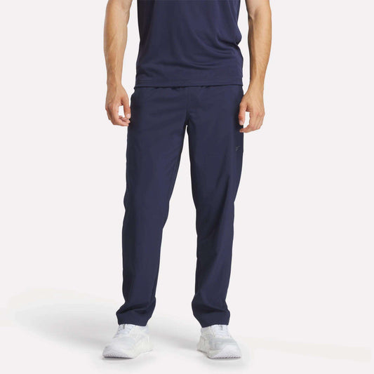 A person stands facing forward against a plain light background, wearing Reebok Men's Train Woven Unlined Pants in navy blue with a matching t-shirt and white athletic shoes. The person's head is not visible in the image.