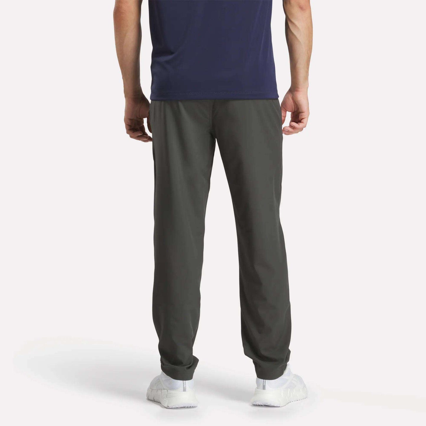 A person stands facing away, wearing a navy T-shirt, Reebok Men's Train Woven Unlined Pants ideal for gym sessions, and white sneakers against a plain white background.