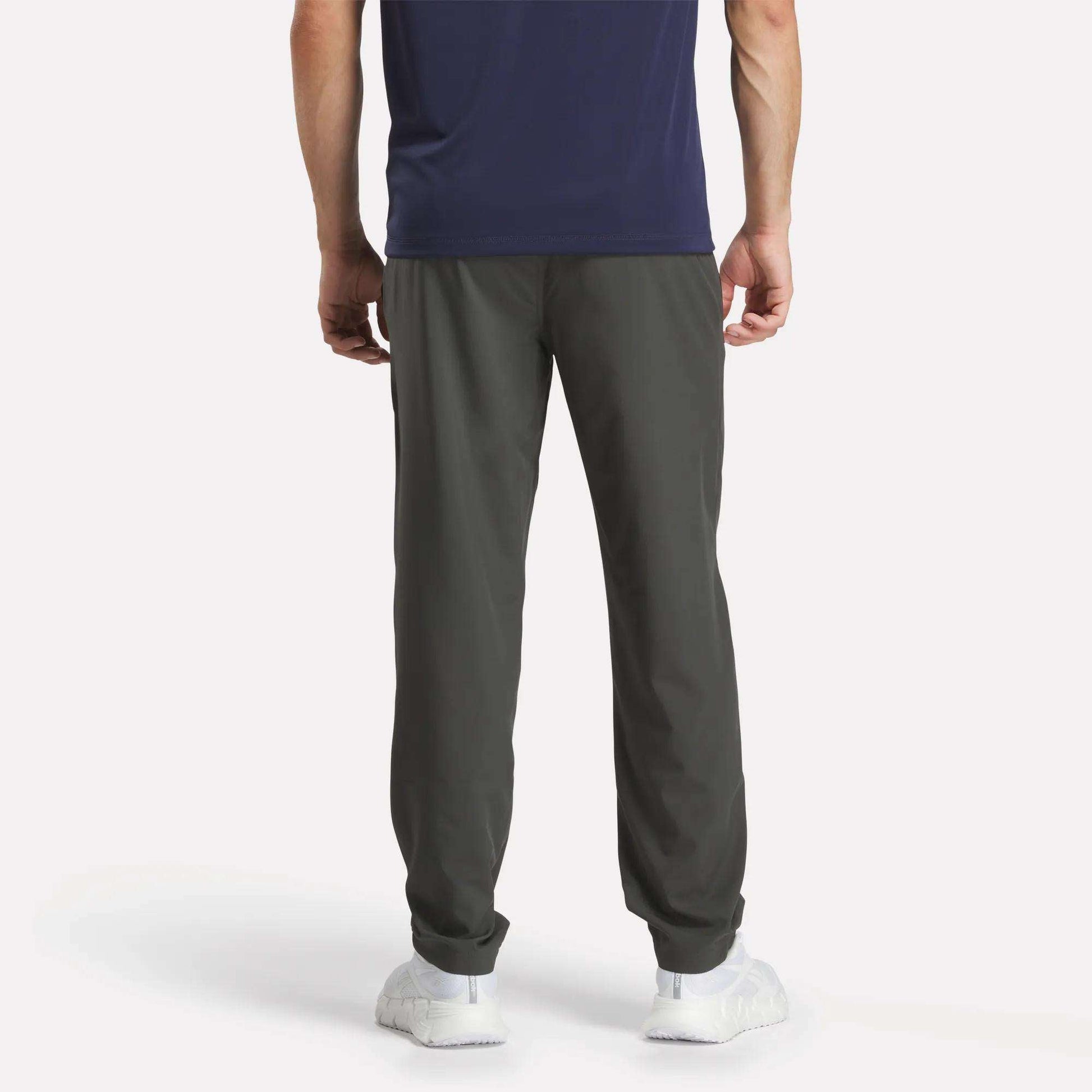 A person stands facing away, wearing a navy T-shirt, Reebok Men's Train Woven Unlined Pants ideal for gym sessions, and white sneakers against a plain white background.