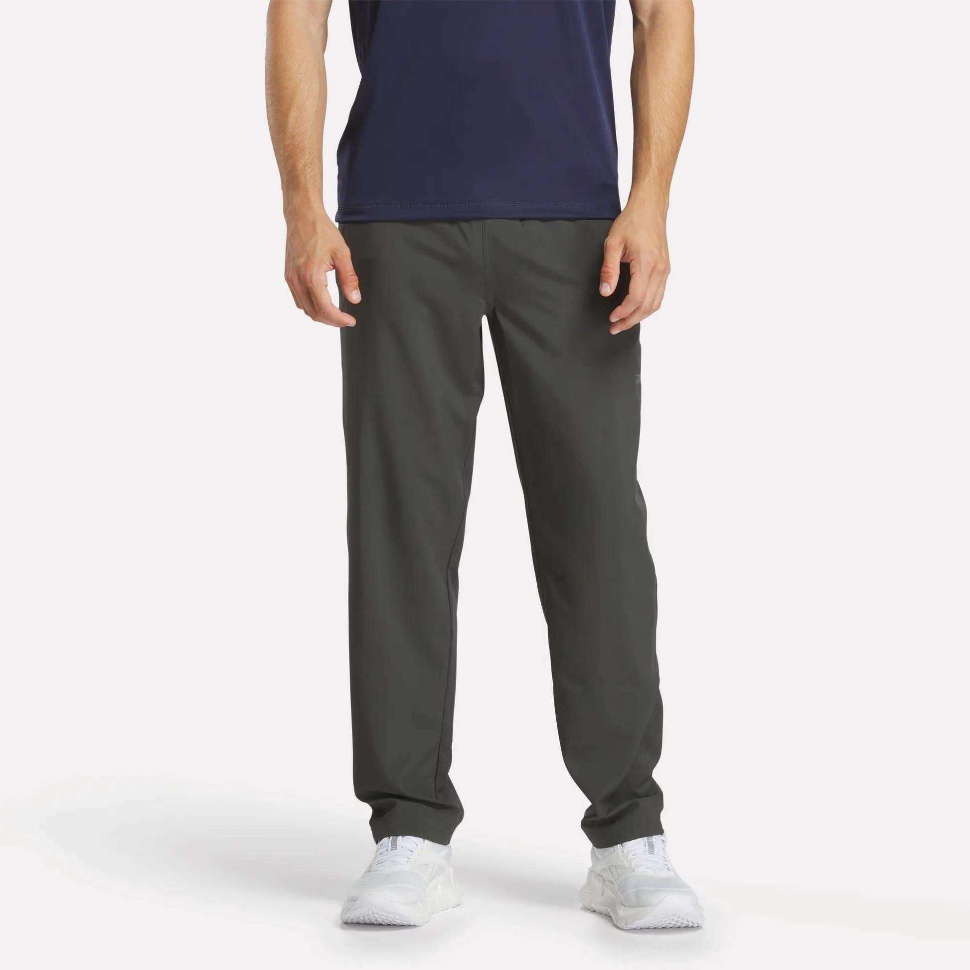 A man stands facing forward against a plain white background wearing Reebok Men's Train Woven Unlined Pants, a navy shirt, and white sneakers. The image shows him from shoulders to feet—ideal attire for gym workouts.